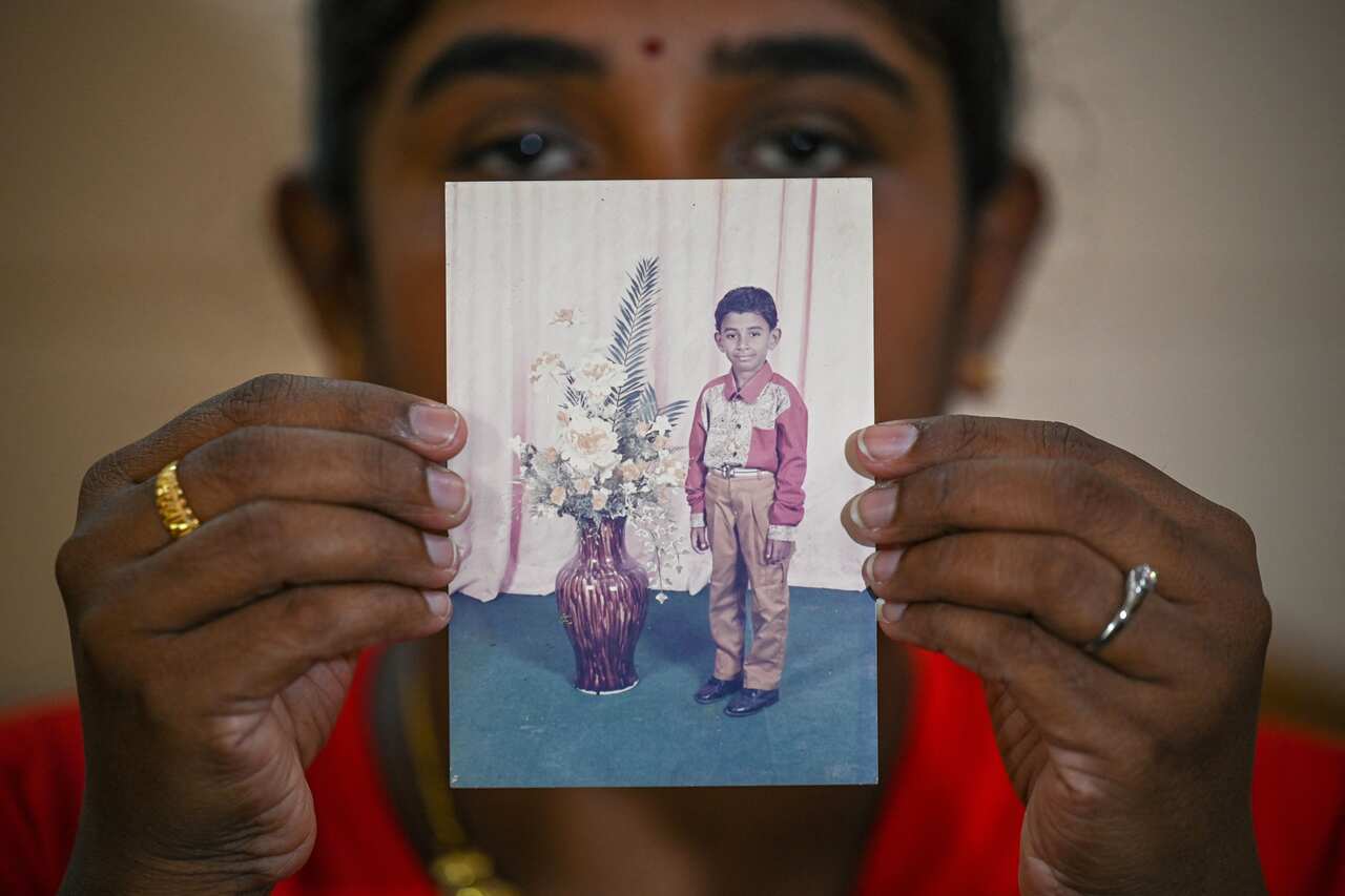 Woman in background carries a portrait photograph of a young boy standing near a large vase of flowers. 