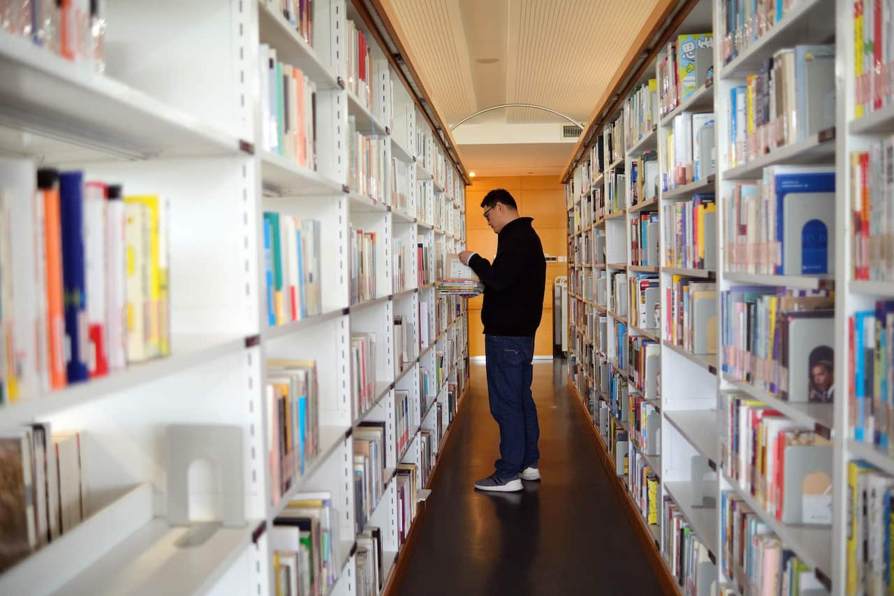 A man wearing a black knit and pants stands between white shelves filled with books inside a library.