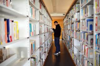 A man wearing a black knit and pants stands between white shelves filled with books inside a library.