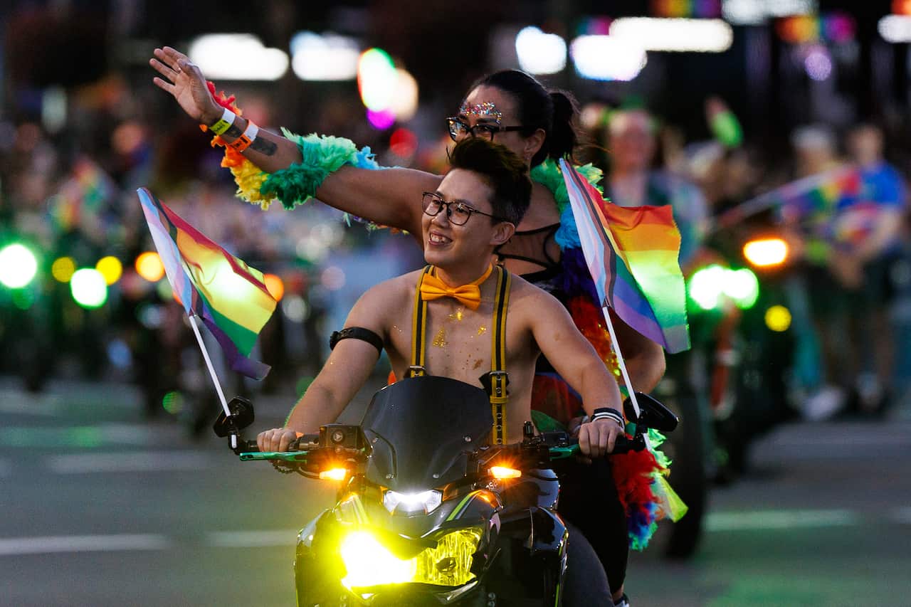 A joyful person with glasses and a yellow bow tie rides a motorcycle in a night parade. Two Progress Pride flags are mounted to the handlebars. Behind them, a passenger with glitter on their face waves to the crowd, draped in a voluminous, multi-colored feather boa.