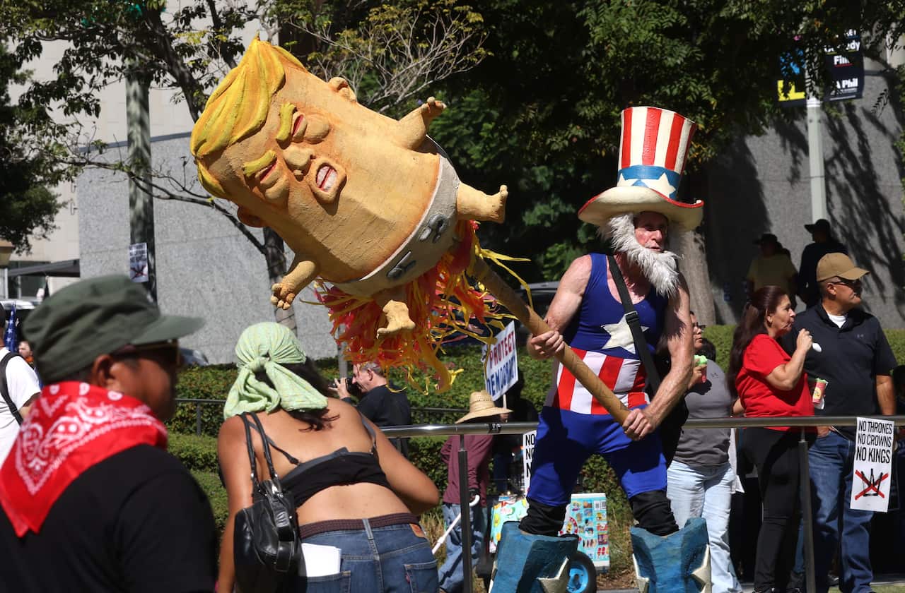 A costumed protester dressed as Uncle Sam, complete with a starred and striped top hat, tank top, and blue boots, swings a large stick at an enormous, grotesque papier-mâché effigy of Donald Trump.