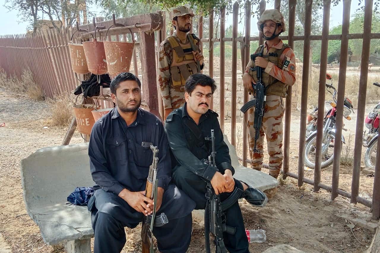 Two people sitting with guns, with two security personnel with guns standing behind them