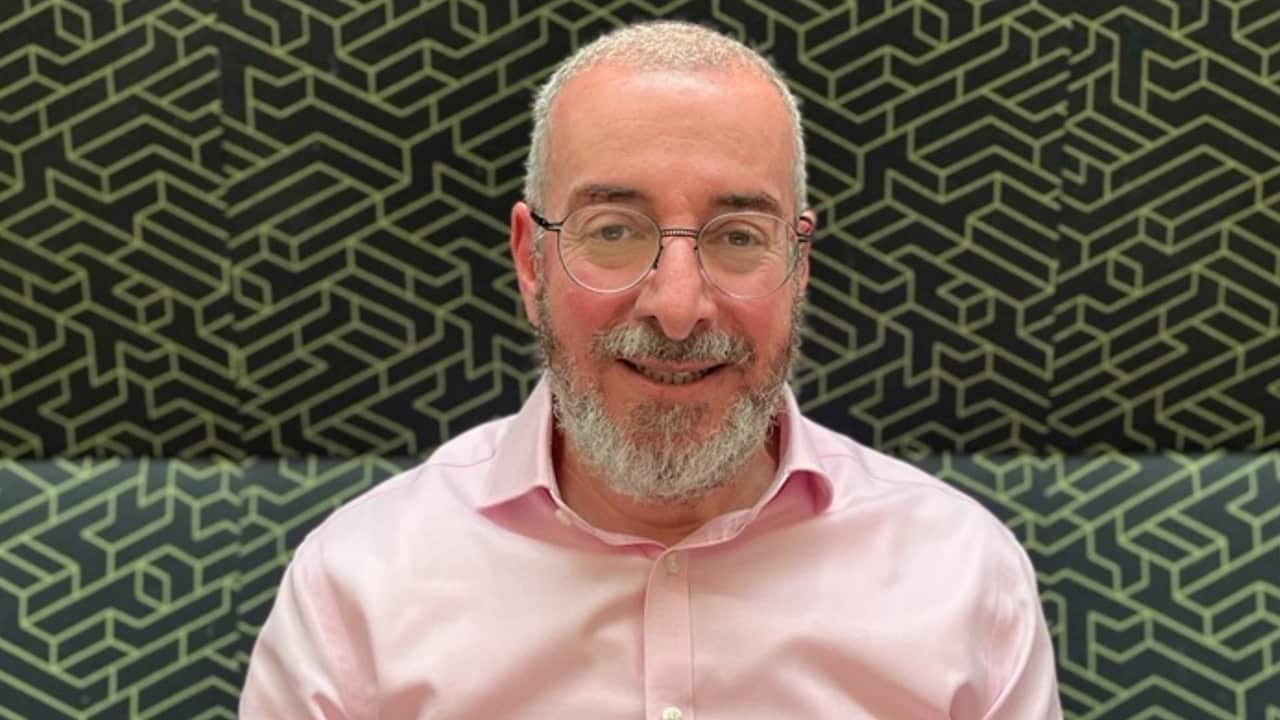 closeup of a man with a grey beard and short-cropped hair. He is wearing a pink shirt and sitting on a high-backed couch