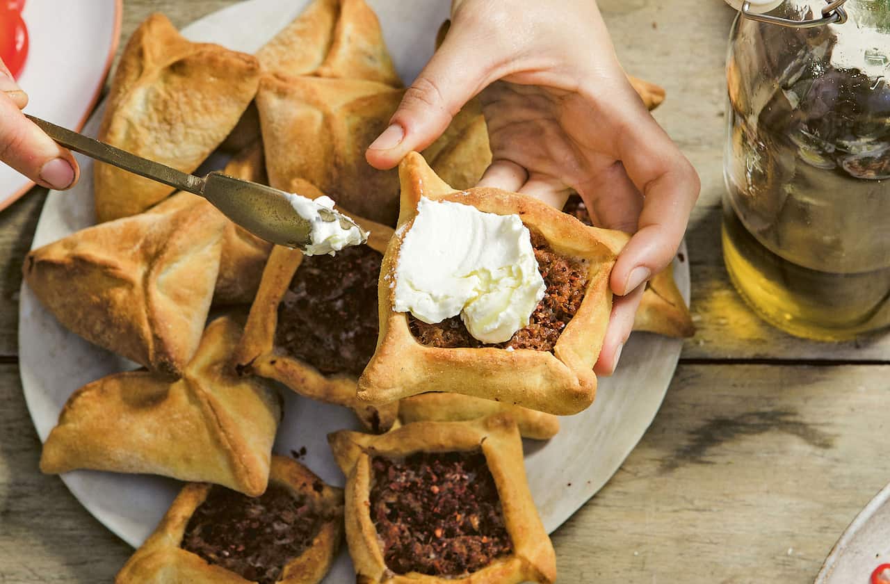 A hand holds a square pastry above a plate, topping it with labna. 