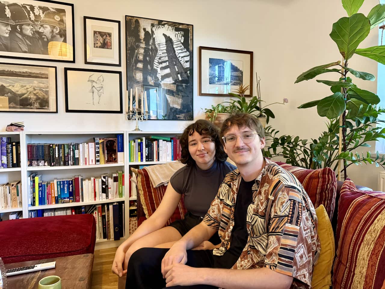 A smiling young couple sits on a patterned red sofa in a cozy room filled with framed art, a large fiddle-leaf fig plant, and white bookshelves packed with colourful books.