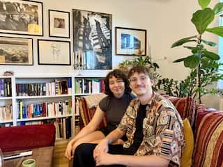 A smiling young couple sits on a patterned red sofa in a cozy room filled with framed art, a large fiddle-leaf fig plant, and white bookshelves packed with colourful books.