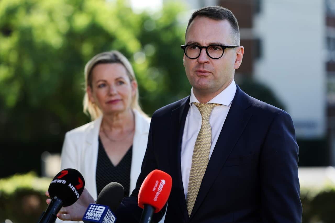Woman watches man in suits speaking in front of microphones.
