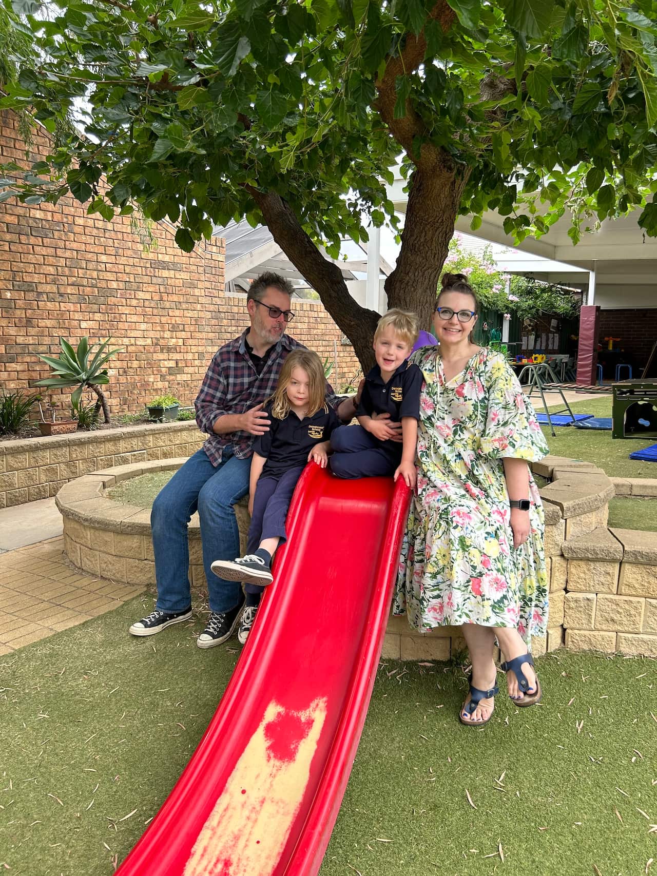 A couple with their two children in a rear lawn. One of the little boys is sitting on a red slide.