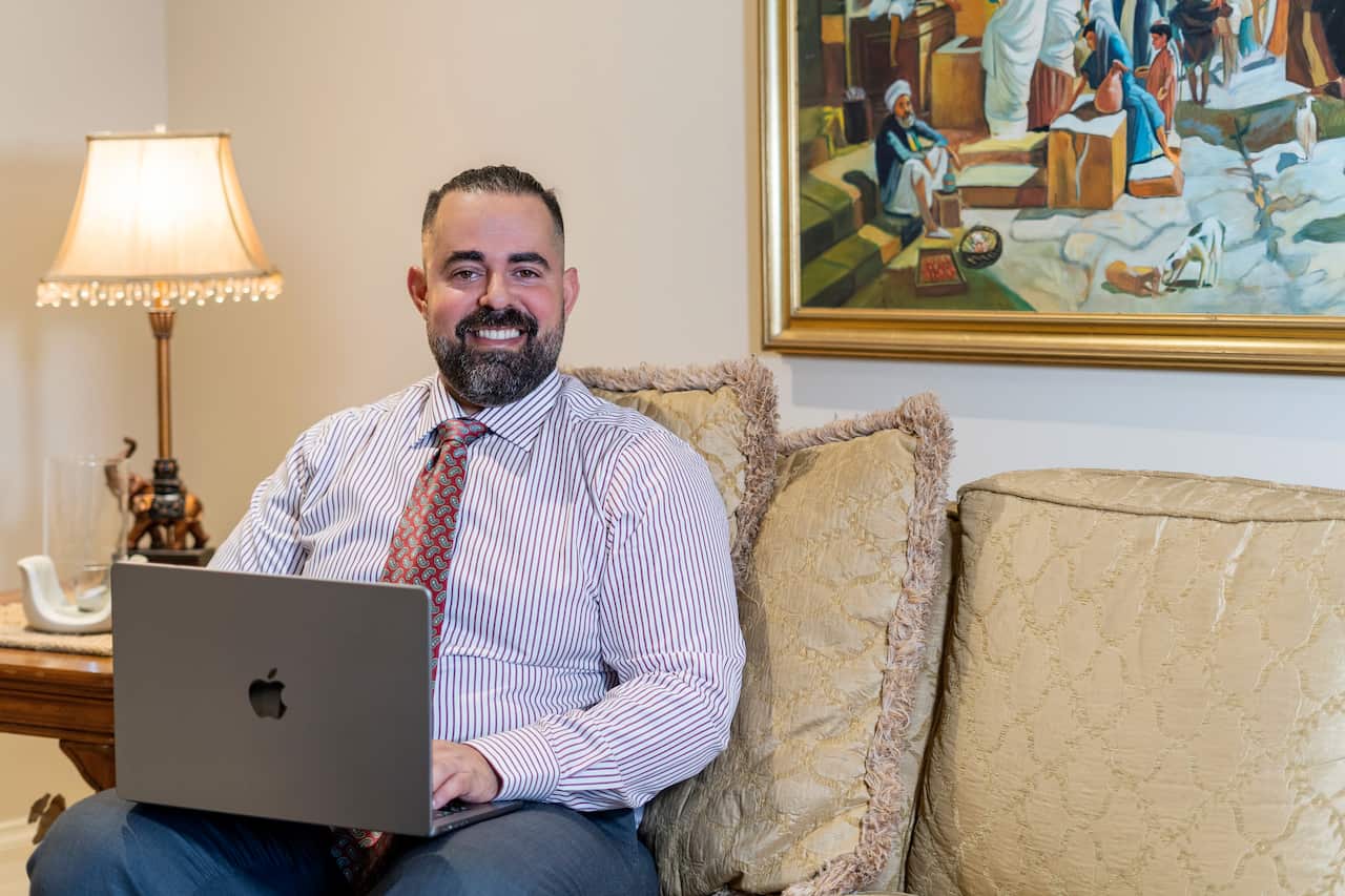 A man sits on a couch with a laptop, smiling in a room decorated with a lamp and a framed painting.