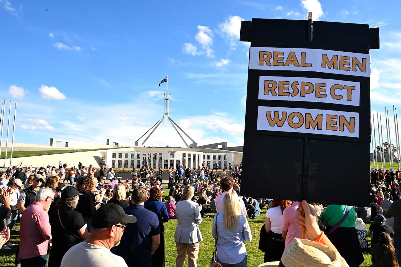 A large group of people standing on the lawn in front of Parliament House in Canberra. A person in the foreground is holding a sign reading: "Real men respect women".