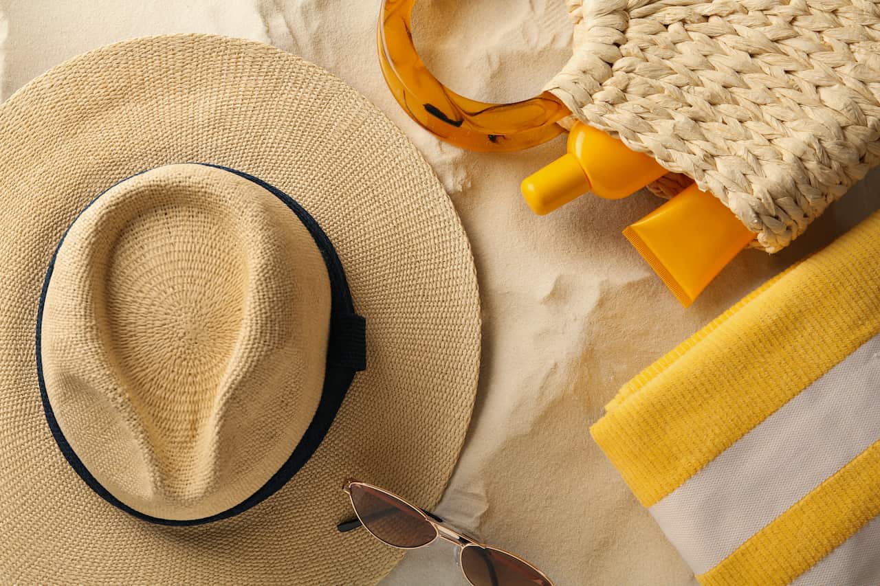 A hat, sunglasses, towel and bag with sunscreen in it on the sand.