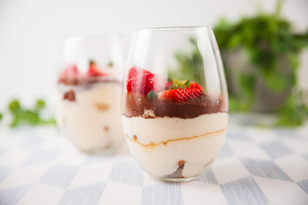 Creamy layers half-fill an elegant bulb-shaped glass. Chocolate and fresh berries sit on top of the creamy tiramisu. Another filled glass can be seen, out of focus, behind the first. The pair of glasses sit on a pale checkered tablecloth. 