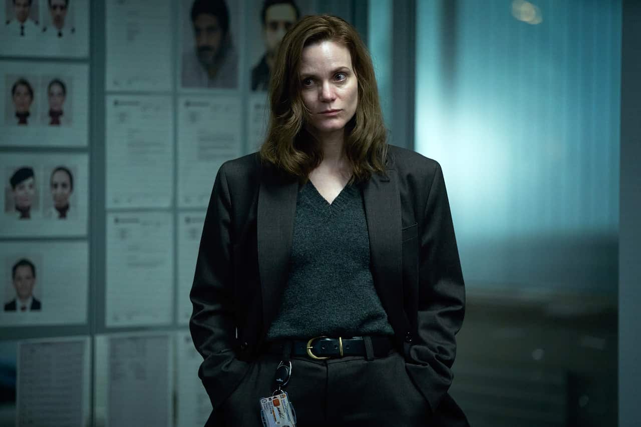 A woman in a blazer stands in a dark room. Printed photographs and other pages are lined up on the wall behind her.