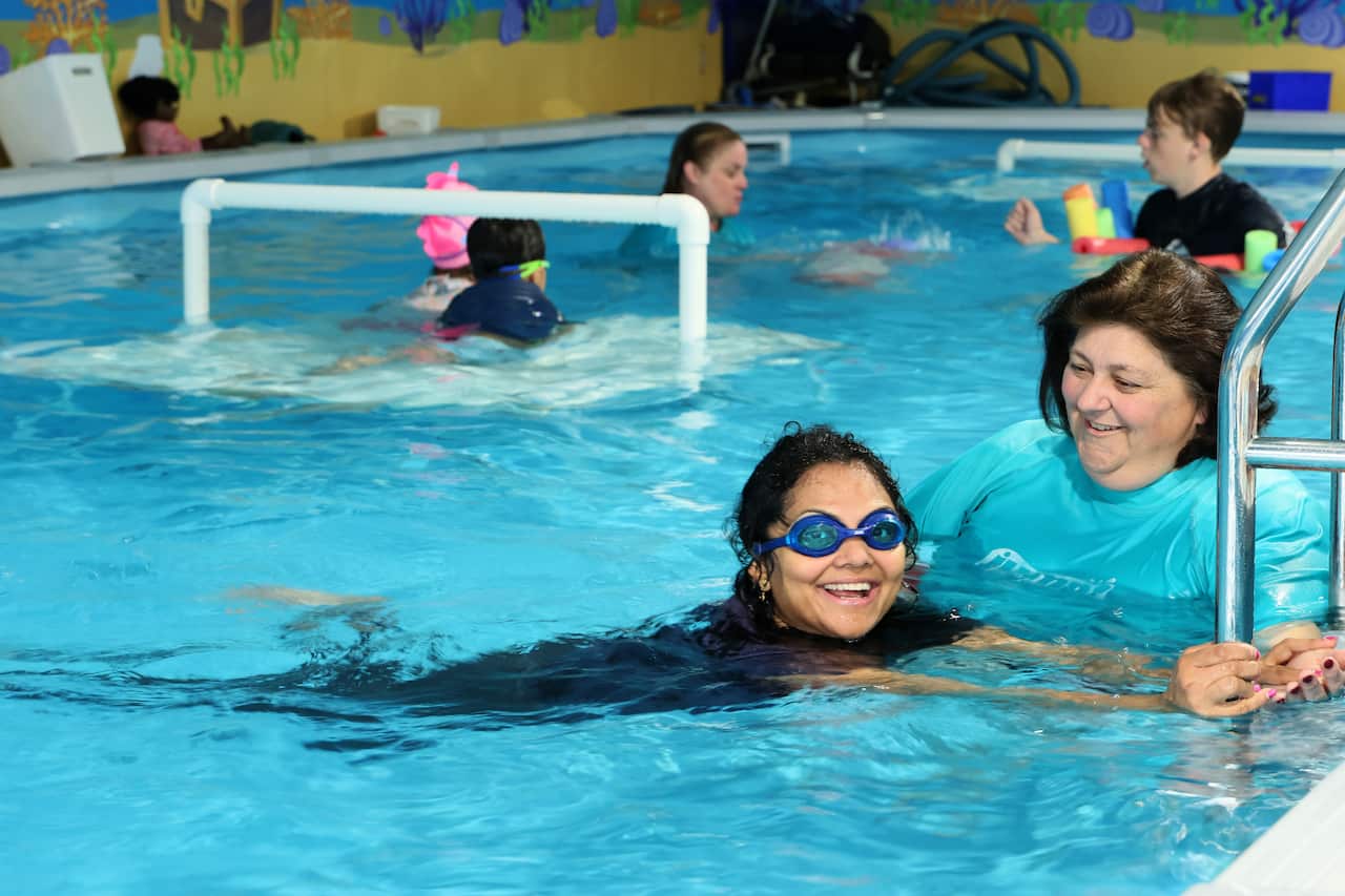 A woman in a swimming pool holds onto the railing and smiles. A swimming instructor is supporting her.