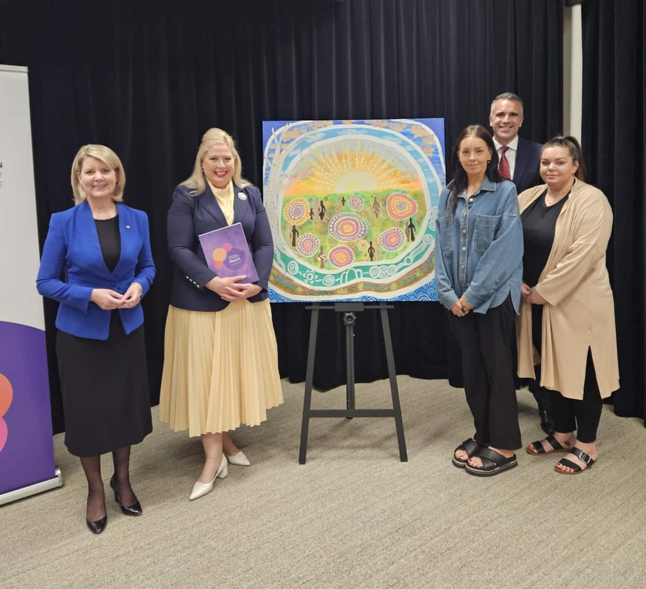 A woman on the left in a blue jacket, a woman second to the left in a cream skirt, a painting on a canvas in the centre, two women on the right with a man in a suit behind them.