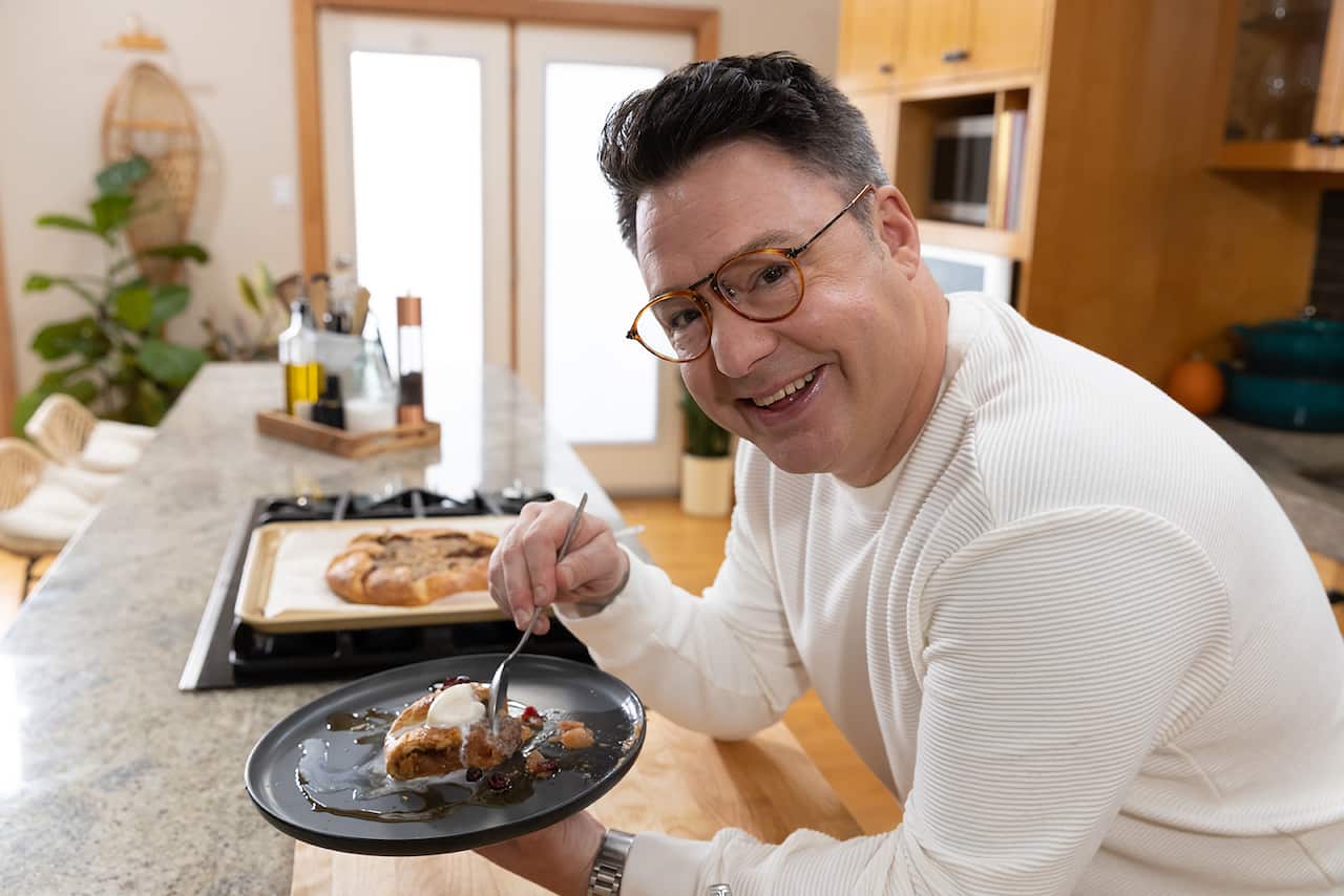 A man in a white long-sleev knit top sits side on in a kitchen, holding a plate with a slice of something sweet drizzled with cream. He has a happy smile on his face. 
