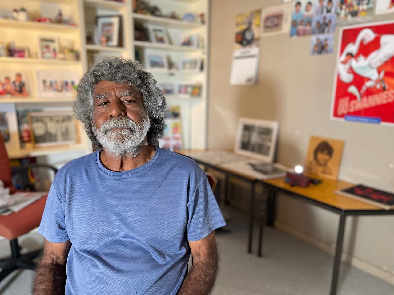 Gomeroi man Don Craigie sits in the loungeroom of his Tamworth home in a blue t-shirt, surrounded by photos of his family. He has grey curly hair and a grey beard, and is looking solemn, directly to camera.