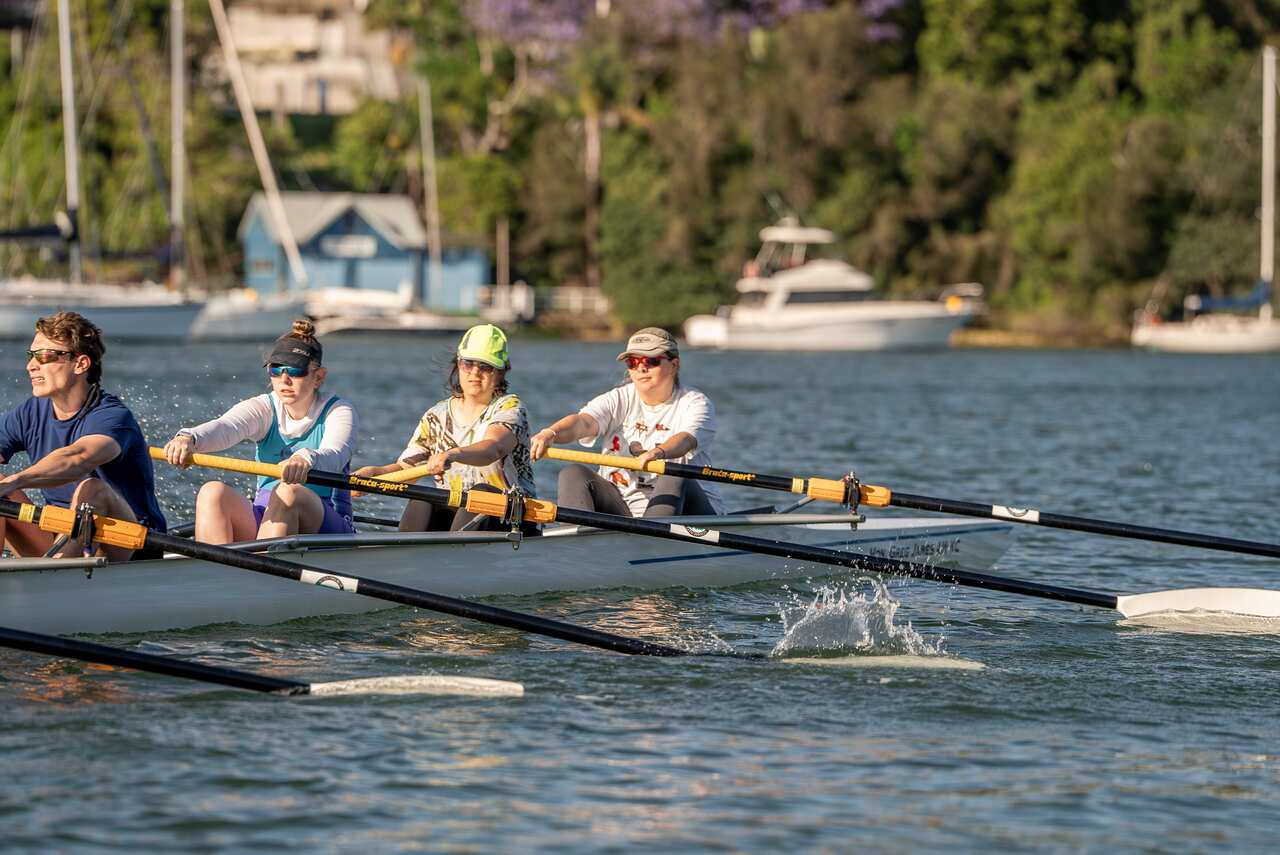 Four rowers, each holding a single oar, row in unison in a boat on the water, with other boats visible in the background.