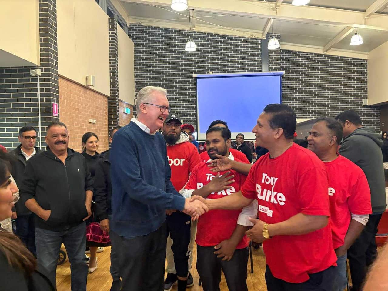 A man in a suit shakes hands with a person in a red t-shirt in front of a crowd of other people wearing red t-shirts 