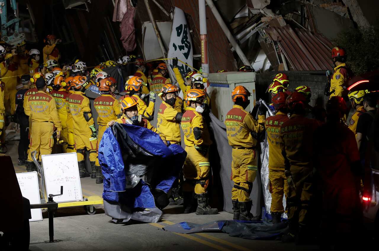 A large group of people in yellow jumpsuit uniforms stand amid the wreckage of buildings. 