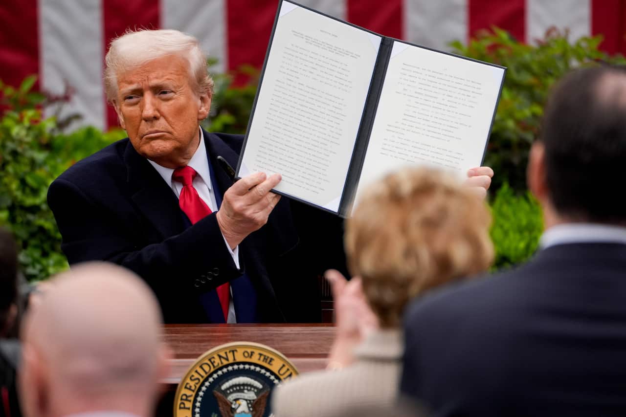 Donald Trump wearing a suit standing at a podium, holding up an executive order in front of a crowd
