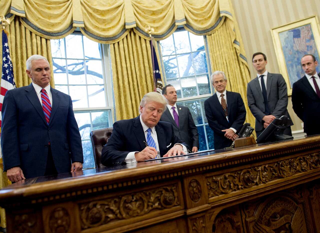 Donald Trump signing papers in the White House while men around him watch.