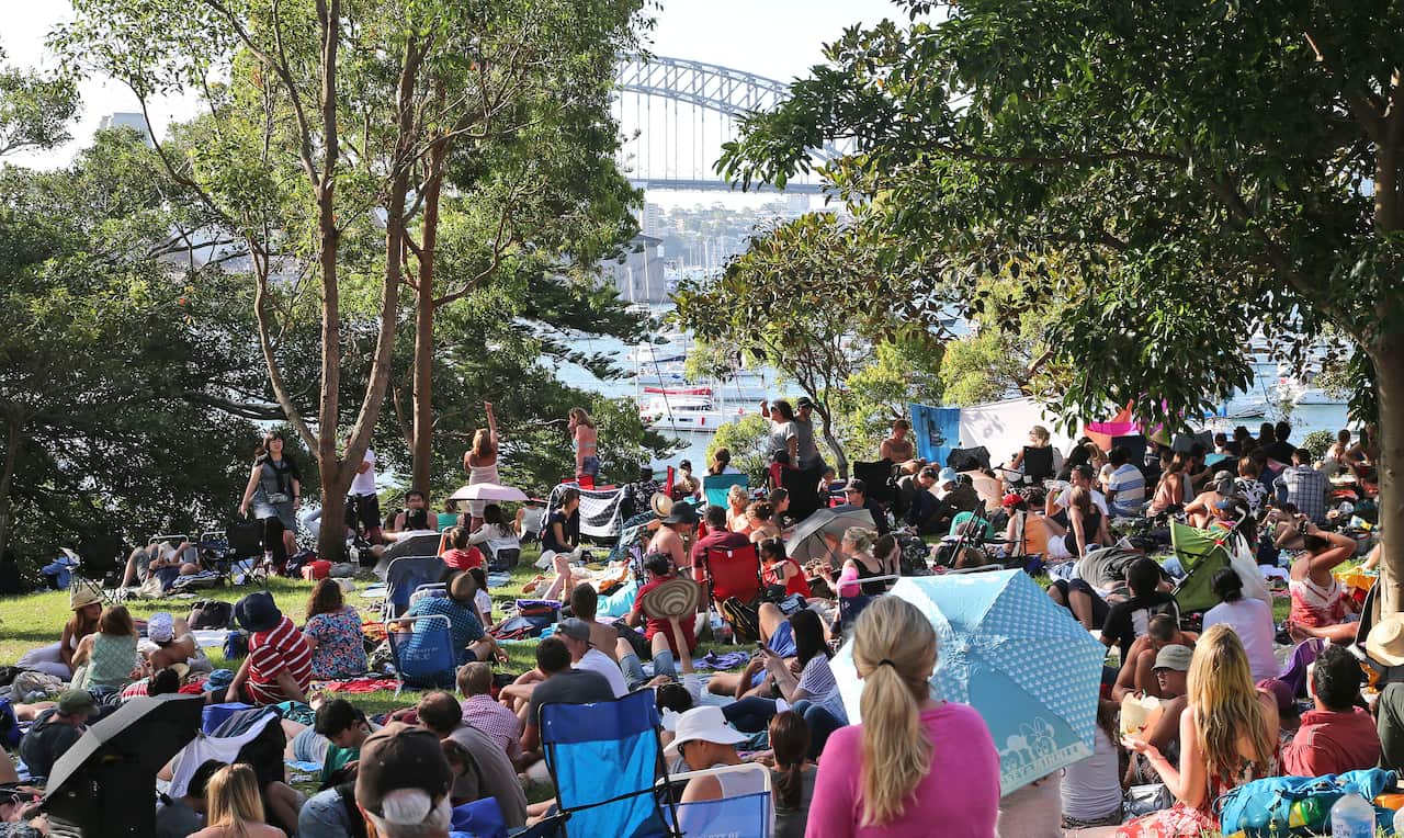 A crowd of people lay on the grass and on picnic blankets in a park overlooking the Sydney Harbour Bridge and the ocean.