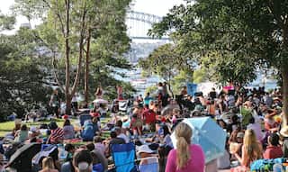 A crowd of people lay on the grass and on picnic blankets in a park overlooking the Sydney Harbour Bridge and the ocean.