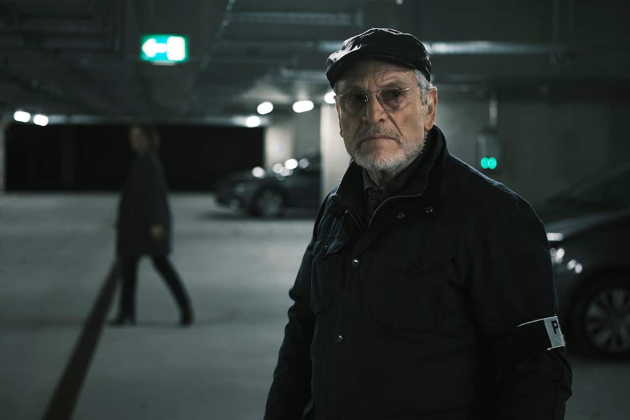 A man in a dark coat and cap and glasses, with a small white beard, stands in an underground carpark. A figure can be seen walking in the distance. 