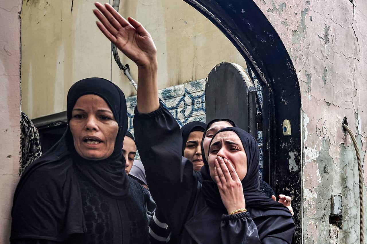 Women cry as they stand at a doorway. One has her hand raised.