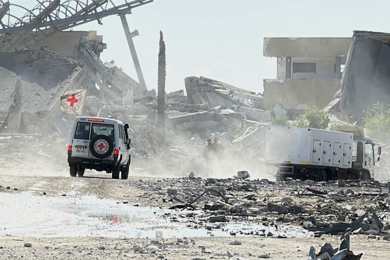 A Red Cross vehicle passes through a dirt road flanked by destroyed buildings and rubble. There's a white truck ahead.