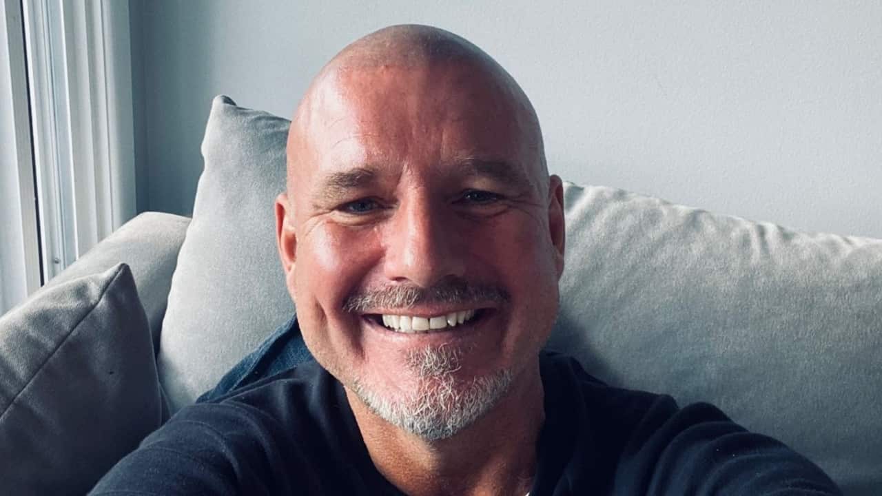 a middle-aged man with a shaved head and beard stubble smiles widely while taking a selfie as he sits on a couch