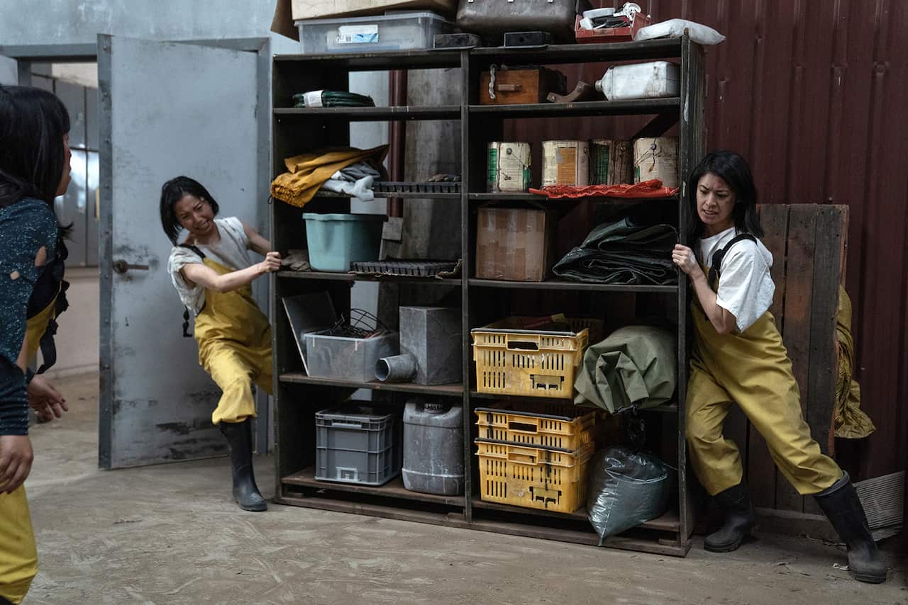 Two women in yellow overalls and black gumboots desperately try to move a shelf, while a third can be seen just on the edge of the image, and looks to be saying something urgent. 