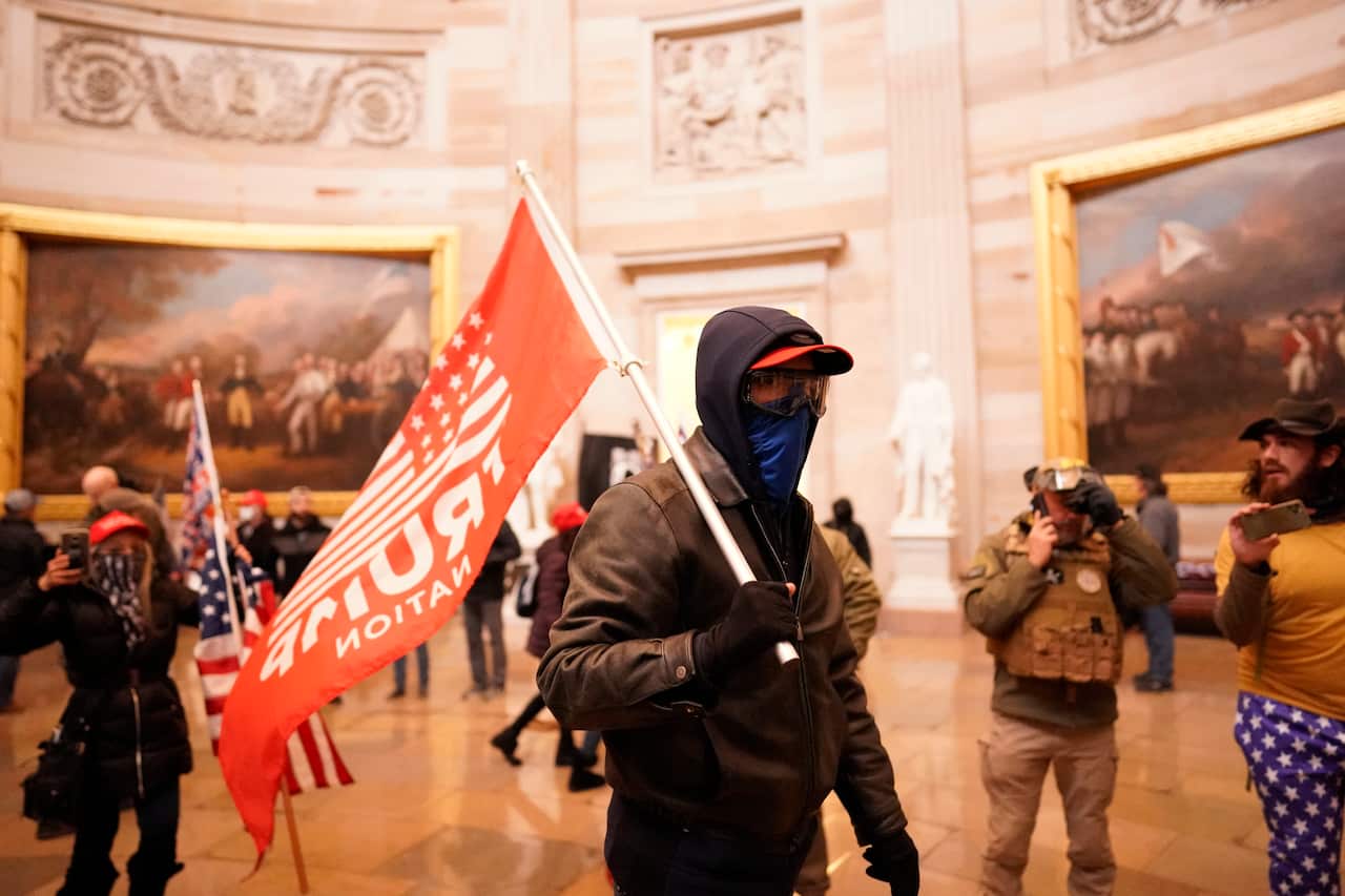 A man in a mask holds a red flag with the word Trump on it inside a historical-looking building.