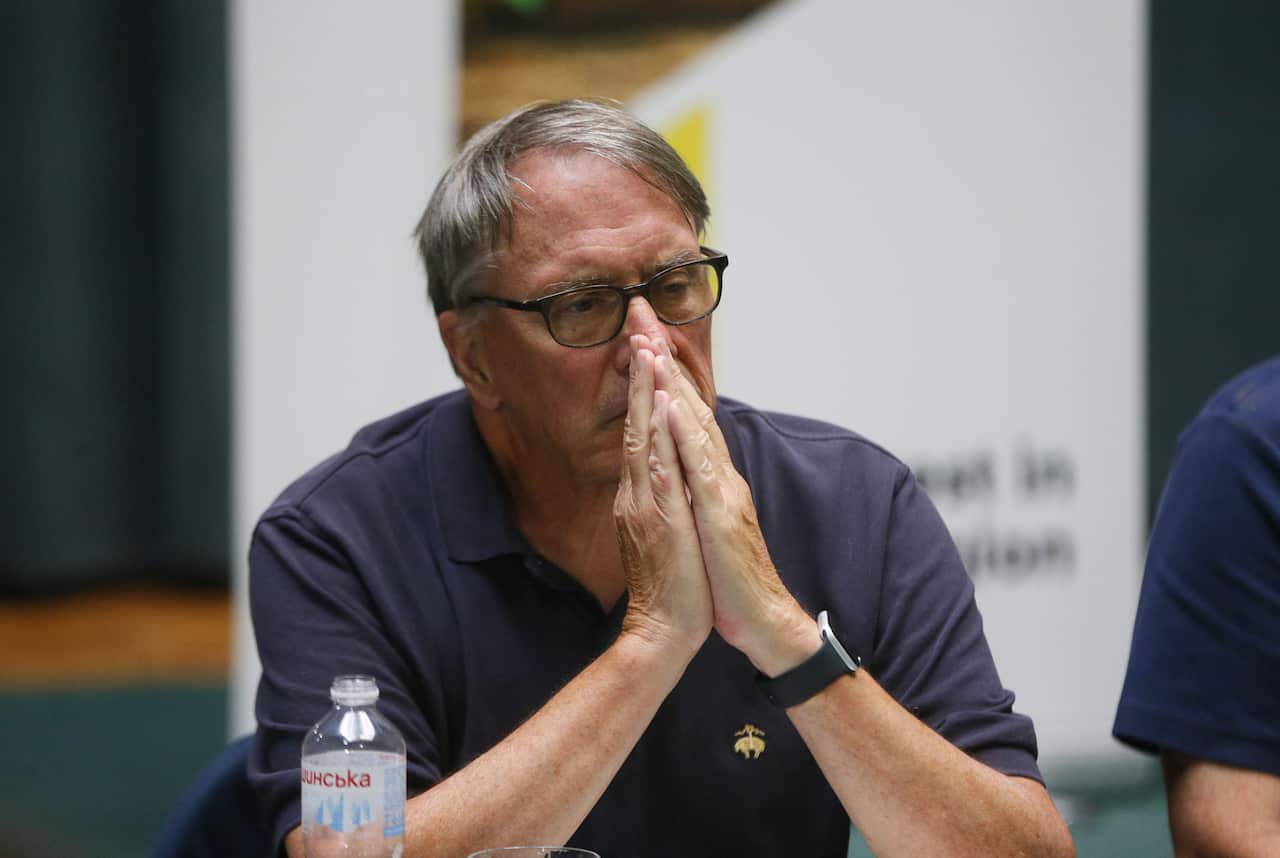 A man wearing glasses and a dark blue polo shirt sits with his hands held clasped in front of his face.