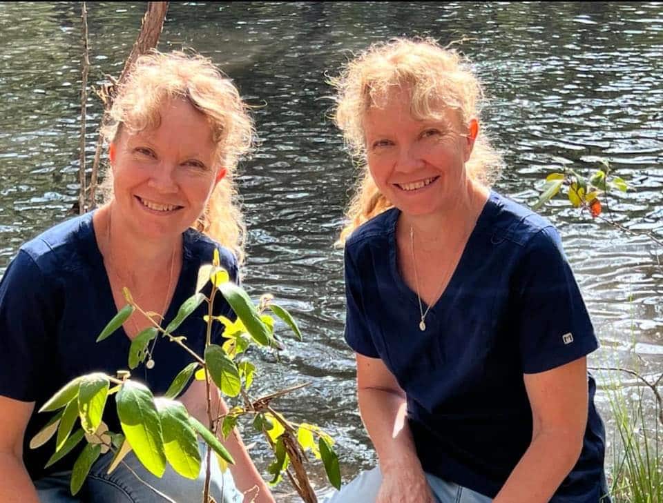 A pair of identical twins wearing identical blue shirts kneel behind a sapling. There is a lake in the background. 
