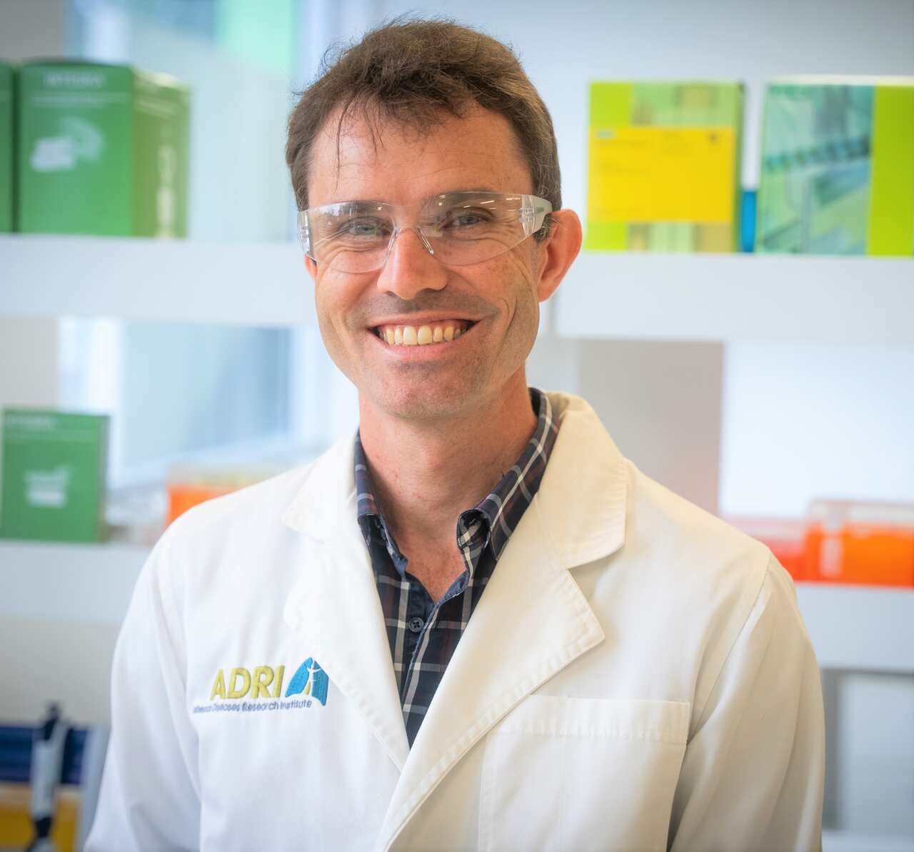 A smiling man wearing a white lab coat and clear protective glasses.  