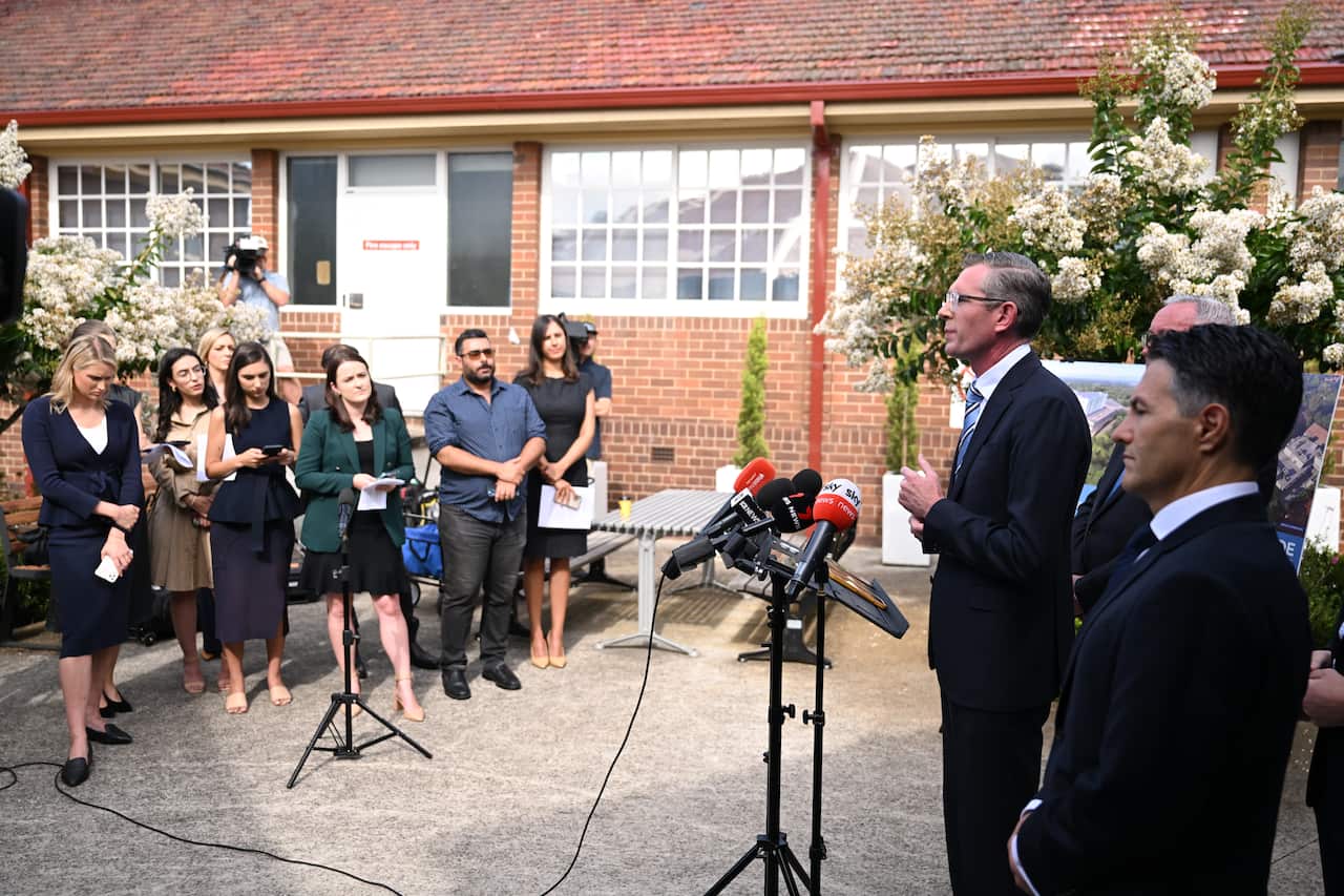 A group of journalists watch NSW Premier Dominic Perrottet who is speaking in front of a microphone.