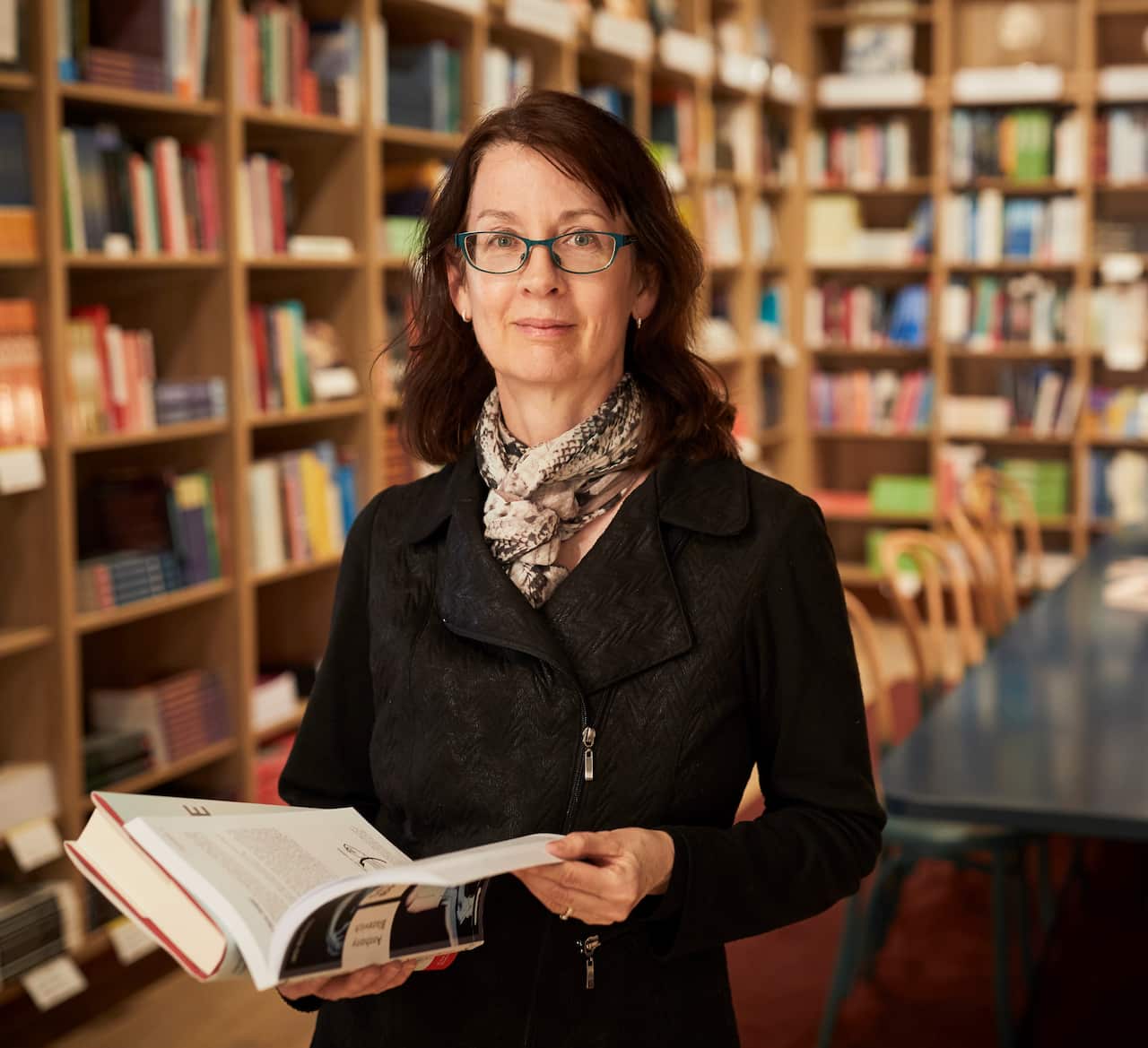 A woman wearing glasses stands in a library, holding a book open