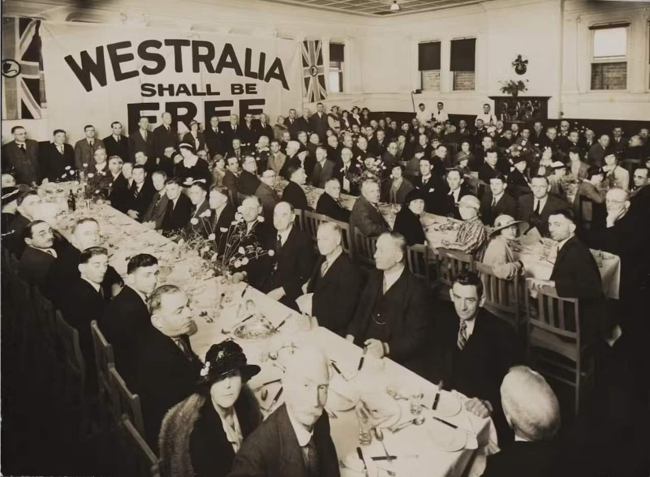 A black and white image of people sitting at tables next to the sign 'Westralia shall be free'. 