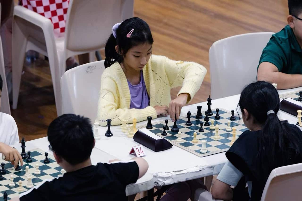 A young girl wearing a pale yellow cardigan is playing chess against another young girl.
