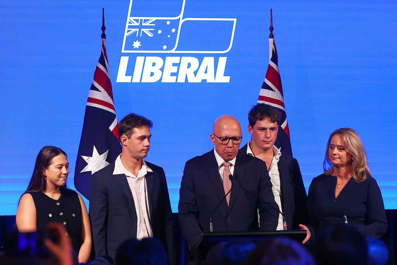 A bald man stands at a lectern looking down, surrounded by four other people. There are two Australian flags and a blue backdrop with a Liberal logo behind them
