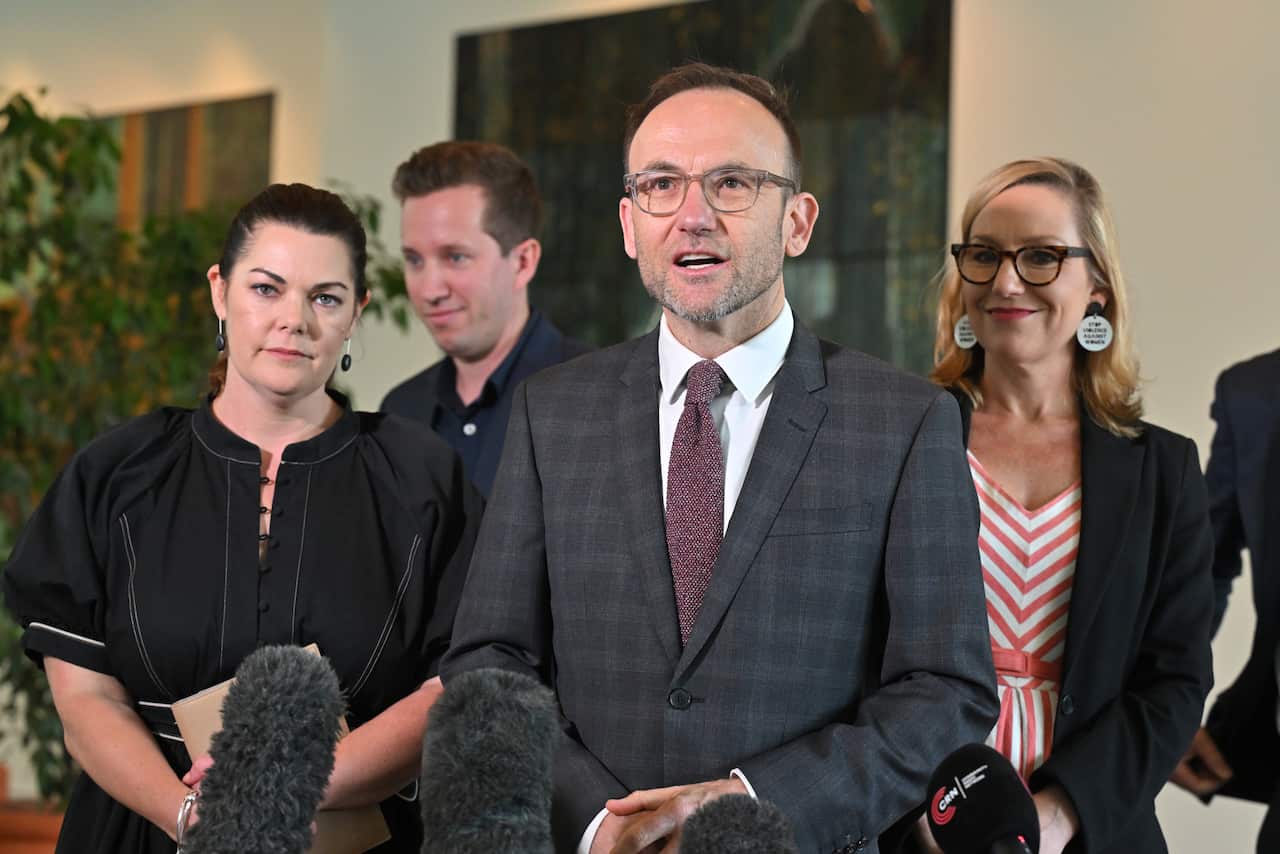 A man in a dark grey tweed suit speaks, looking ahead of him with two women on either side.