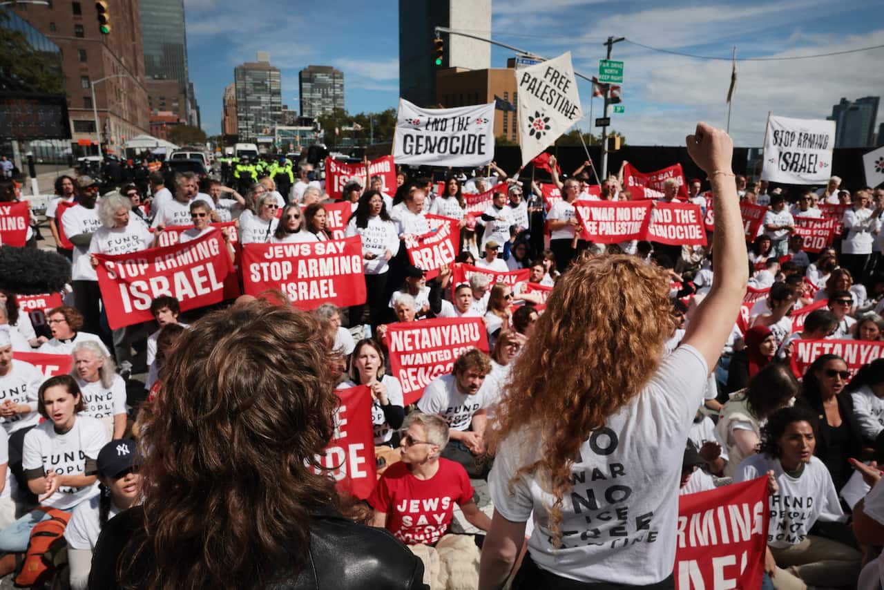 A crowd of protestors holding red banners calling to "end the genocide" and "stop arming Israel".