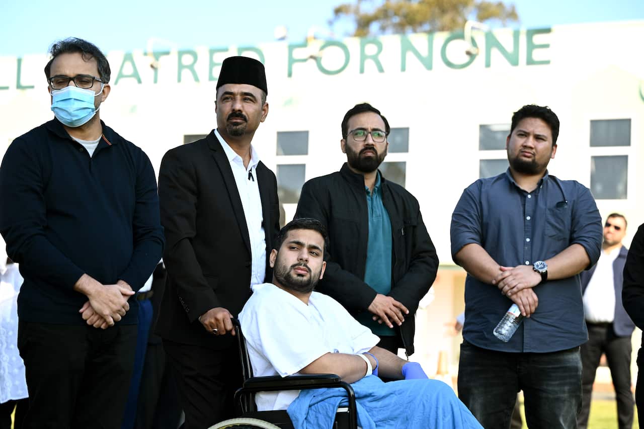 A man in a wheelchair and a hospital gown, with four men wearing black standing behind him.