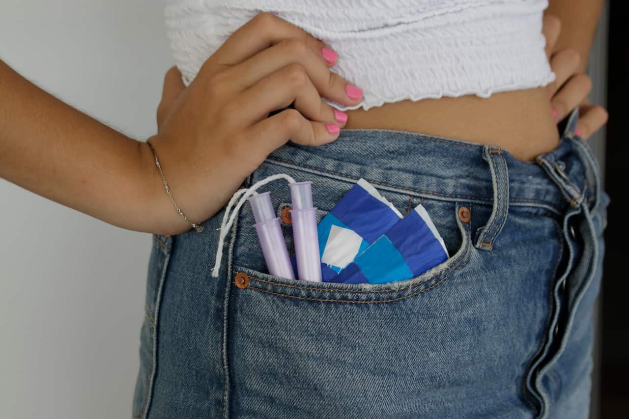 The midsection of a woman's body with her hands on her waist. Tampons and pads are spilling out of her jeans pocket.