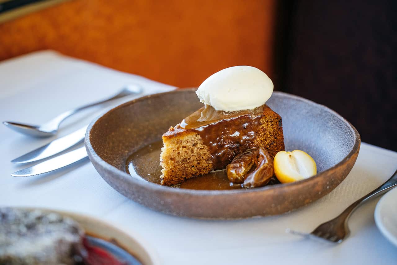 A wedge of cake-like sticky date pudding sits in a stylish rounds bowl, topped with a gleaming caramel coloured sauce and a scoop of ice-cream