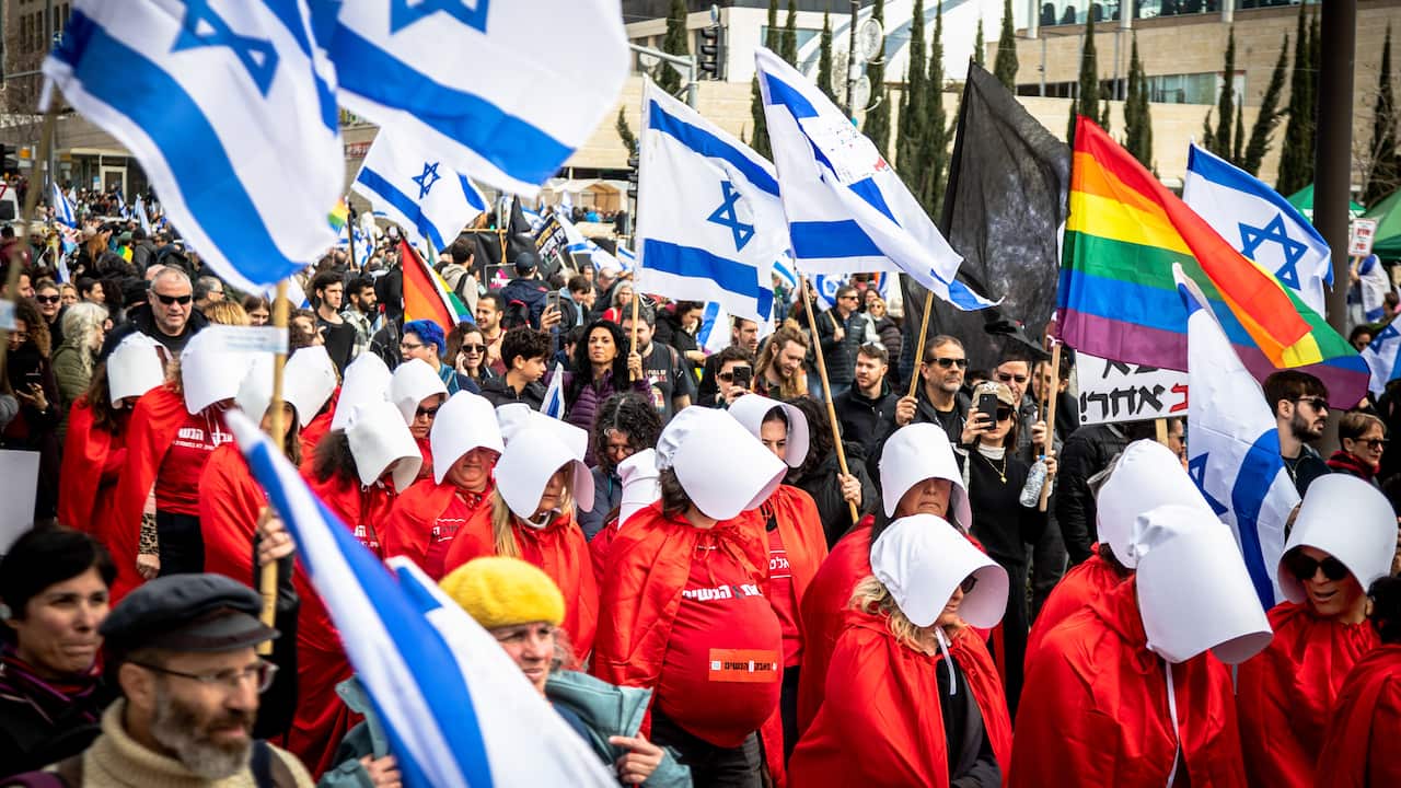 People dressed in Handmaid's Tail costumes marching in a protest. Some people are carrying Israel flags on poles and there is also a rainbow flag.