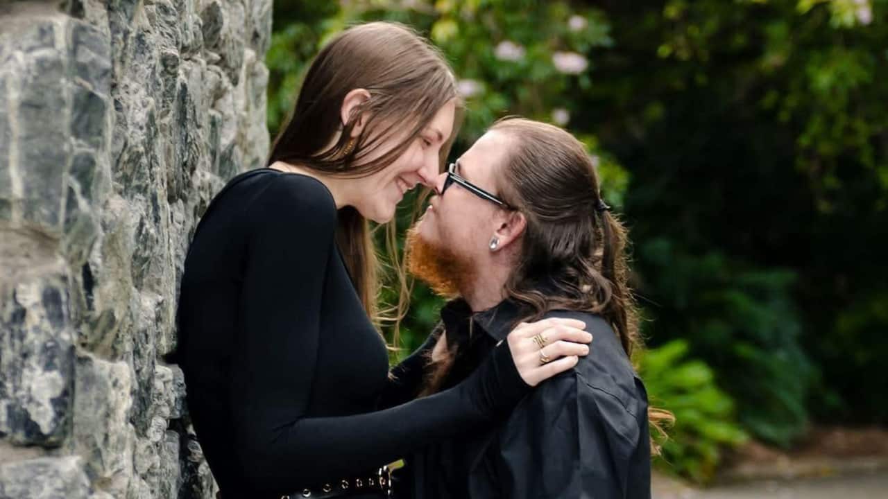 a tall young woman stands against a tree smiling and touching noses with her shorter boyfriend