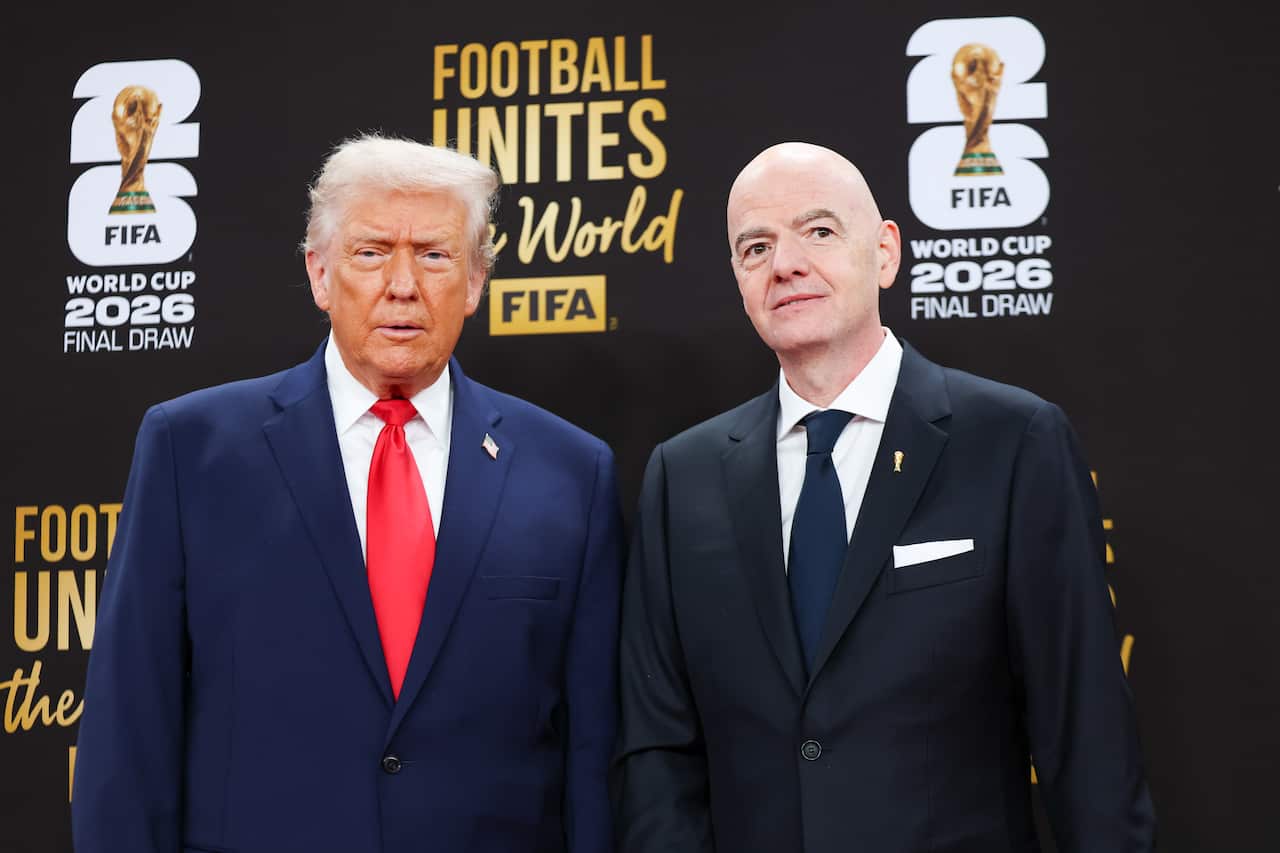 An older white man with blonde hair, next to a middle-aged bald man. Both are wearing suits and standing in front of a sign promoting the World Cup.